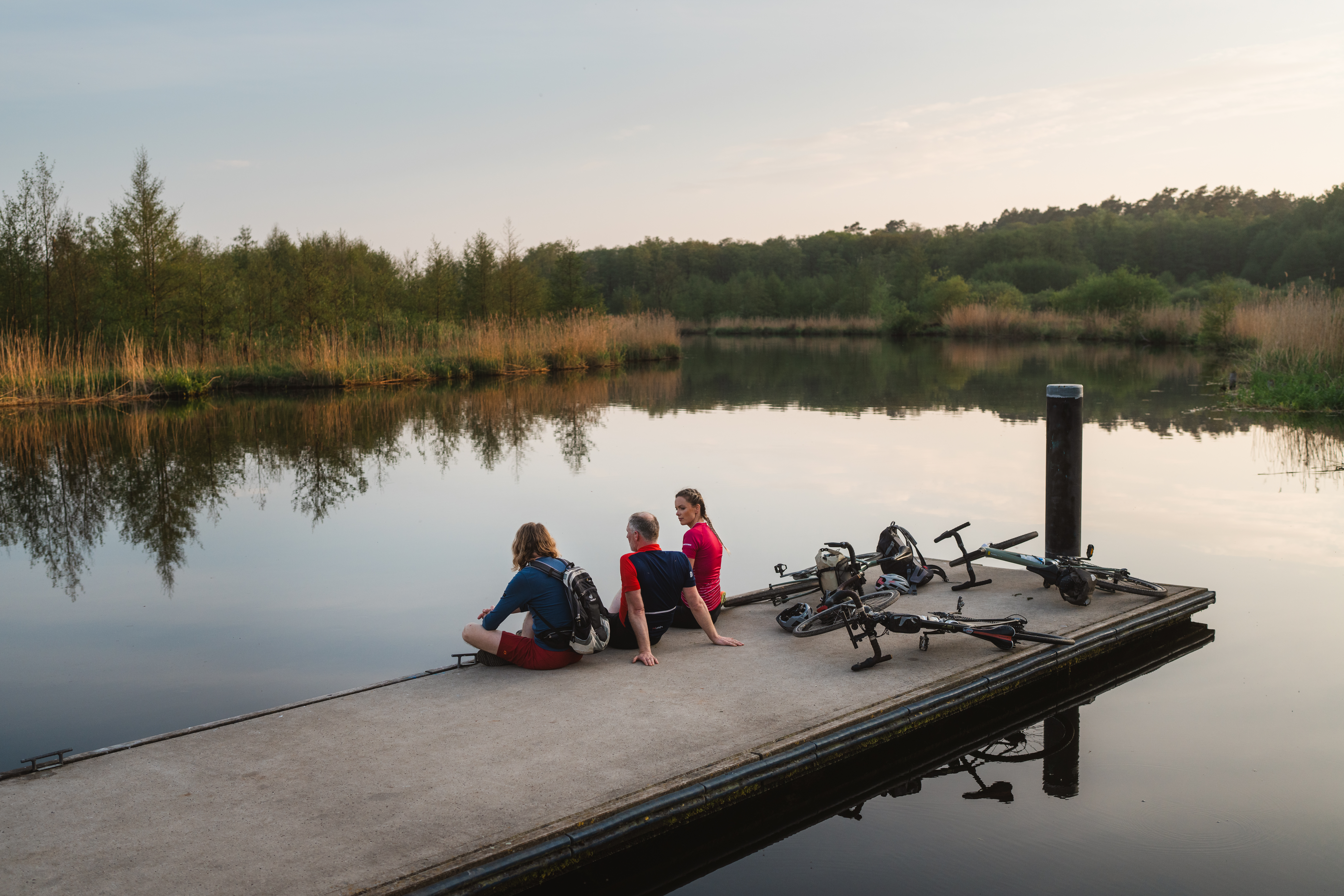 Drei Personen sitzen am Steg an der Peene, den Blick aufs Wasser gerichtet. Hinter ihnen liegen drei Fahrräder. Im Hintergrund sieht man die Natur der Peeneregion